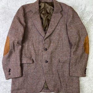 May include: A brown tweed blazer with a classic houndstooth pattern. The blazer features suede elbow patches and three buttons on each cuff. The jacket has a notched lapel and two front pockets. The lining is a solid brown color.