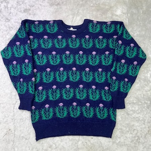 Puede incluir: Un suéter azul marino con un patrón repetido de cardos verdes con cabezas de flores moradas. El suéter tiene cuello redondo y mangas largas. El diseño está tejido en la tela, creando un aspecto texturizado.