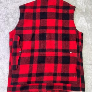 May include: A sleeveless vest featuring a bold red and black buffalo plaid pattern. The garment has a collared neckline, a large rectangular back pocket, and a smaller side pocket secured with a single metal snap button closure on the left side.