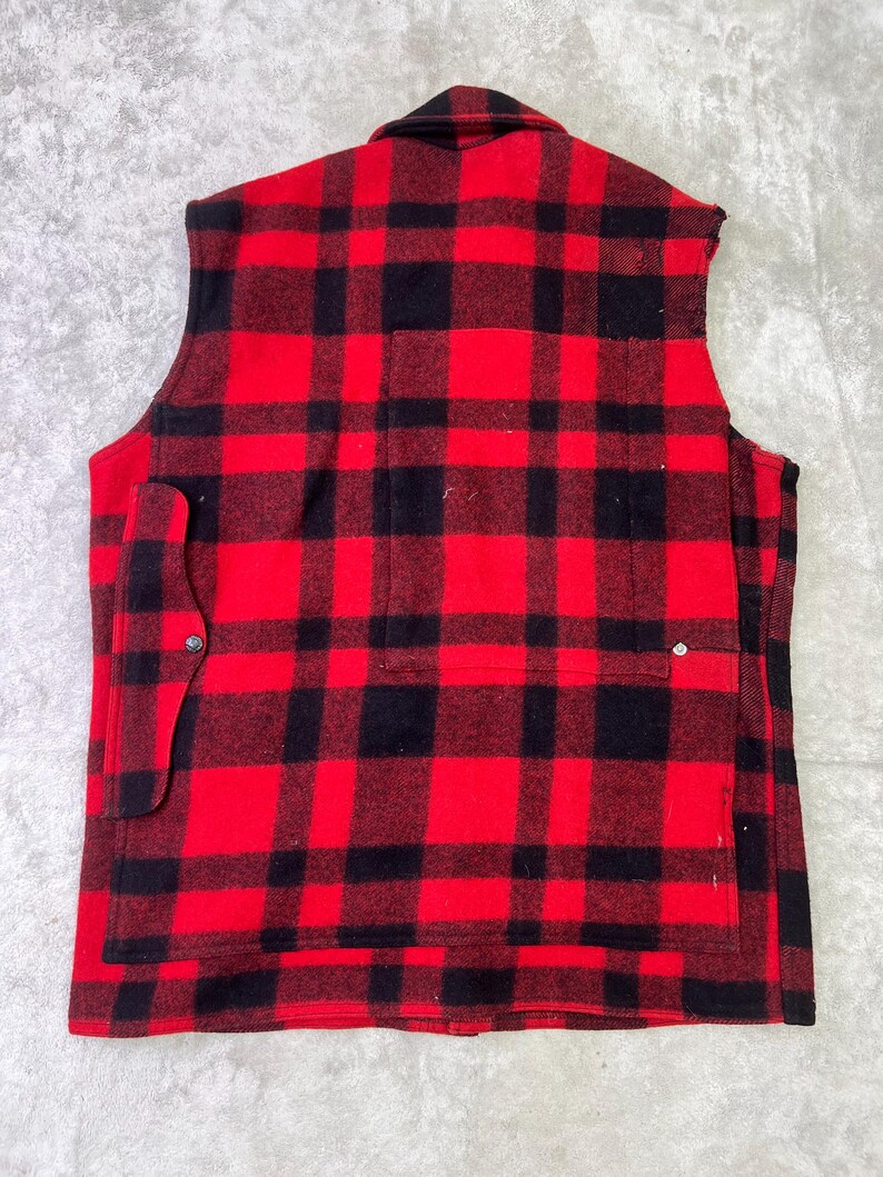 May include: A sleeveless vest featuring a bold red and black buffalo plaid pattern. The garment has a collared neckline, a large rectangular back pocket, and a smaller side pocket secured with a single metal snap button closure on the left side.