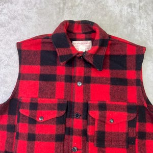 May include: A sleeveless vest in a red and black buffalo plaid pattern. The garment features a fold-over collar, a front button closure, and two large patch pockets on the chest with buttoned flaps. A white label reads Filson Garment.