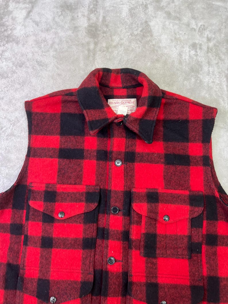 May include: A sleeveless vest in a red and black buffalo plaid pattern. The garment features a fold-over collar, a front button closure, and two large patch pockets on the chest with buttoned flaps. A white label reads Filson Garment.