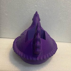 Puede incluir: Un recipiente vibrante en forma de gallina de color p&uacute;rpura. El recipiente tiene un dise&ntilde;o detallado, que incluye un cuerpo texturizado, un borde decorativo en forma de cuerda y una cresta prominente. El cuerpo de la gallina tiene un compartimento abierto.
