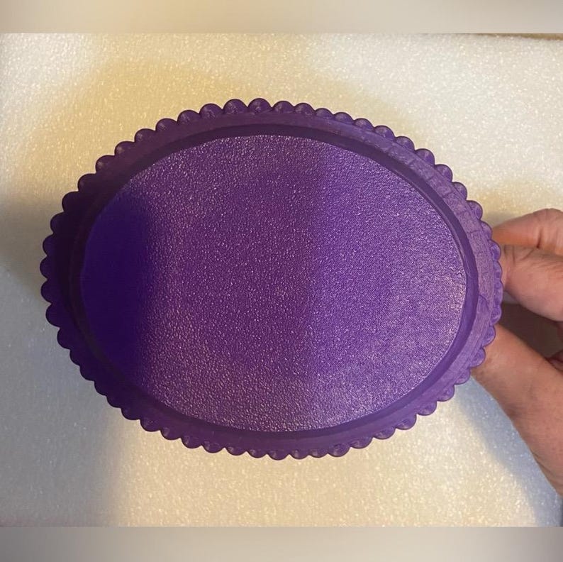 Puede incluir: Una bandeja ovalada morada con una superficie texturizada y un borde festoneado. La bandeja est&aacute; hecha de un material s&oacute;lido y opaco. Una mano sostiene la bandeja.