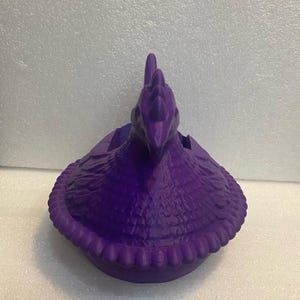 Puede incluir: Un recipiente morado con forma de gallina y superficie texturizada. El recipiente tiene un dise&ntilde;o detallado, incluyendo una cresta y un borde con volantes alrededor de la base. La gallina est&aacute; mirando hacia adelante y parece ser de pl&aacute;stico.