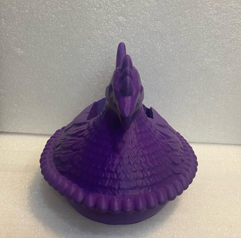 Puede incluir: Un recipiente morado con forma de gallina y superficie texturizada. El recipiente tiene un dise&ntilde;o detallado, incluyendo una cresta y un borde con volantes alrededor de la base. La gallina est&aacute; mirando hacia adelante y parece ser de pl&aacute;stico.