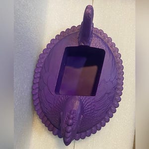 Puede incluir: Un recipiente decorativo morado con un dise&ntilde;o de p&aacute;jaro. El recipiente tiene una abertura rectangular y un borde festoneado. La cabeza y la cola del p&aacute;jaro son caracter&iacute;sticas prominentes, y la textura general parece estar tallada o moldeada.