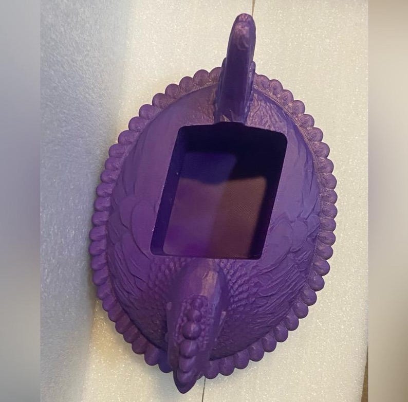 Puede incluir: Un recipiente decorativo morado con un dise&ntilde;o de p&aacute;jaro. El recipiente tiene una abertura rectangular y un borde festoneado. La cabeza y la cola del p&aacute;jaro son caracter&iacute;sticas prominentes, y la textura general parece estar tallada o moldeada.
