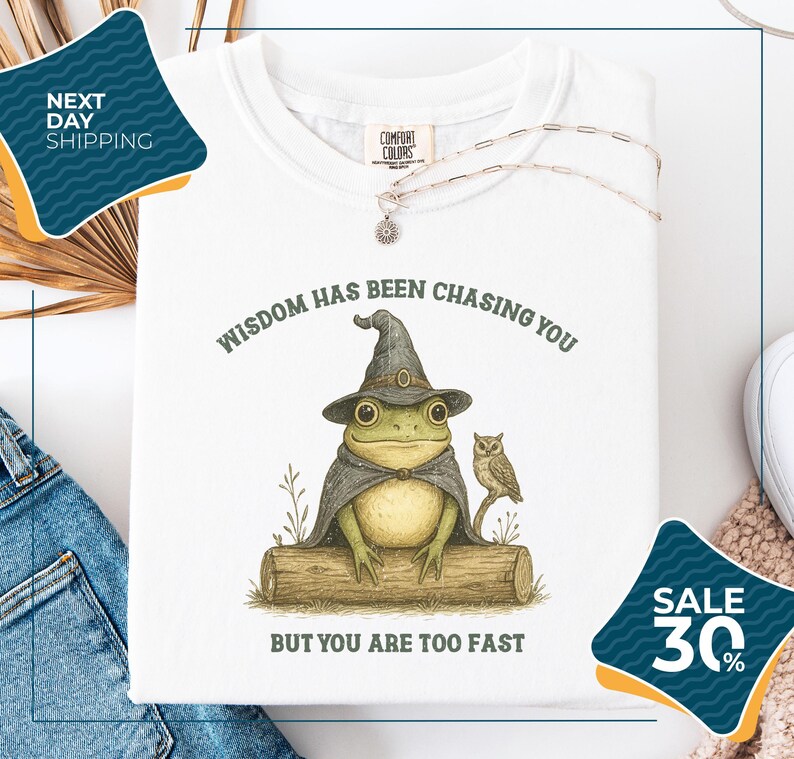 a white t-shirt with a whimsical illustration of a frog wearing a wizard's hat and holding an owl, accompanied by the text "WISDOM HAS BEEN CHASING YOU BUT YOU ARE TOO FAST". The t-shirt is displayed on a white background, with a pair of jeans and a pair of sunglasses visible in the background. There are also two sale signs in the image, one on the left side and one on the right side, indicating a discount or sale event.