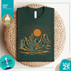 a green t-shirt with a desert landscape design, including cacti and mountains, along with a sun illustration. The t-shirt is displayed on a woven basket, and there are various texts and logos surrounding the image.