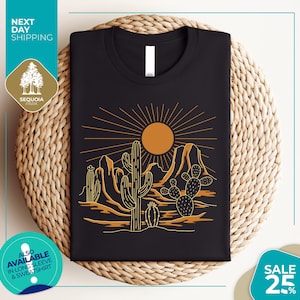 a black t-shirt with a graphic design depicting a desert landscape with cacti and mountains, along with a sun and rays. The t-shirt is displayed on a woven mat, and there are various texts and logos surrounding the image.