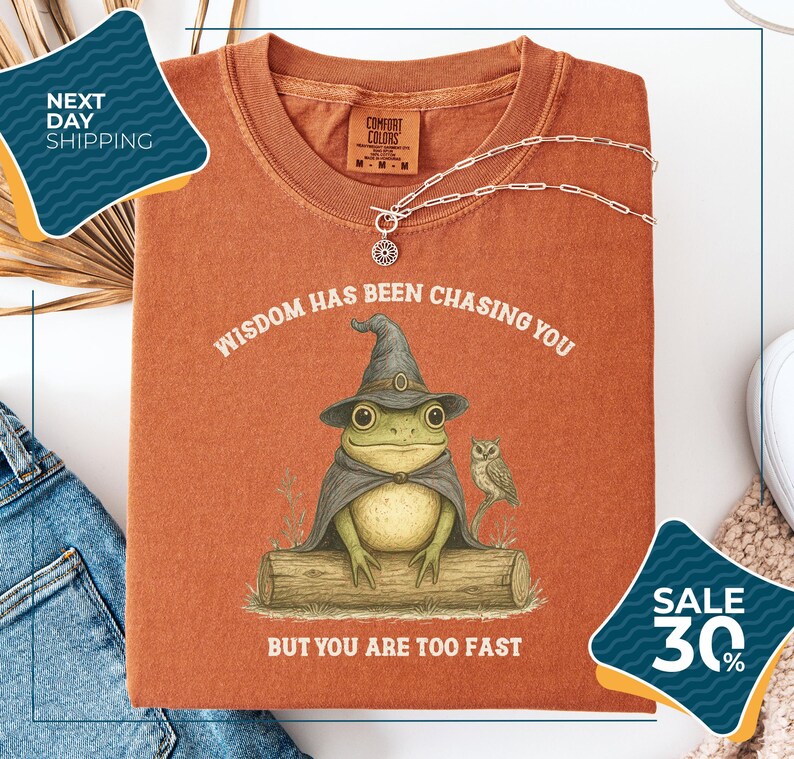 a red t-shirt with a whimsical illustration of a frog wearing a wizard's hat and holding a book, accompanied by the text "WISDOM HAS BEEN CHASING YOU BUT YOU ARE TOO FAST". The t-shirt is displayed on a white background, with a pair of jeans and a necklace visible in the foreground. There are also two sale tags in the image, one for 30% off and another for next day shipping.