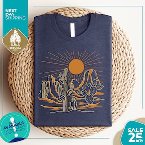 a blue t-shirt with a desert landscape design, including cacti and mountains, along with a sun and rays. The t-shirt is displayed on a woven mat, and there are various texts and logos surrounding the image.