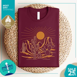 a maroon t-shirt with a desert landscape design, including a sun, cacti, and mountains, displayed on a woven placemat.