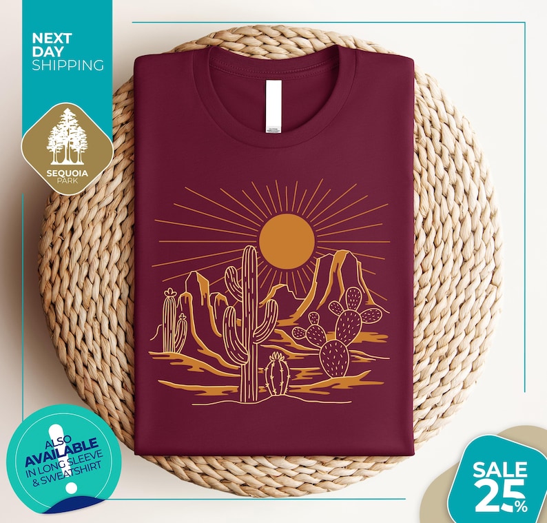 a maroon t-shirt with a desert landscape design, including a sun, cacti, and mountains, displayed on a woven placemat.