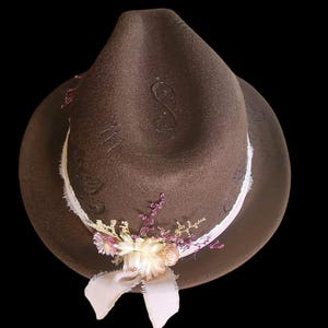 May include: A brown felt fedora hat with a pink ribbon and dried flowers. The hat has a decorative design on the crown.