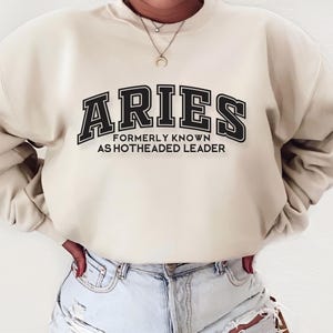 May include: A cream colored sweatshirt with black text that reads "Aries Formerly Known As Hotheaded Leader".