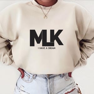 Op de afbeelding: Een crèmekleurig sweatshirt met een zwarte afbeelding van de letters "MLK" en de tekst "I HAVE A DREAM".