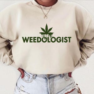 May include: A cream-coloured sweatshirt with a green marijuana leaf graphic and the text "WEEDOLOGIST" printed on the front.