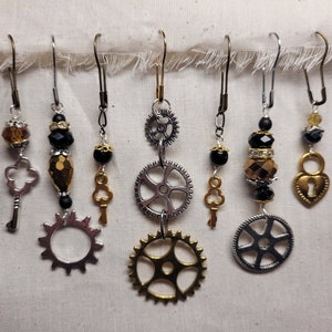 May include: Six pairs of steampunk-style earrings with various charms, including keys, gears, and a heart-shaped lock. The earrings are made with black, gold, and silver beads.
