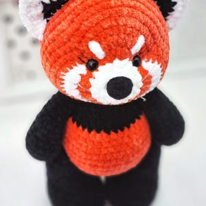 May include: A crocheted red panda plush toy with black and white details. The toy has a red body, black legs and arms, and a white face with black eyes and nose.
