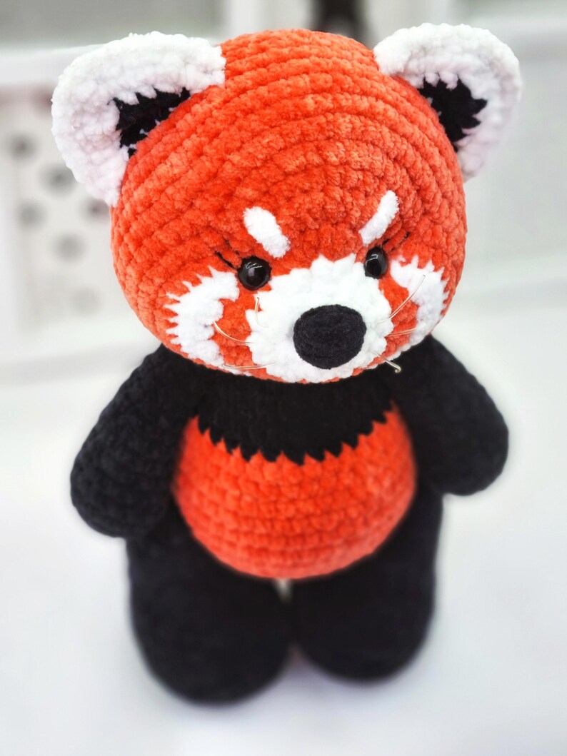May include: A crocheted red panda plush toy with black and white details. The toy has a red body, black legs and arms, and a white face with black eyes and nose.