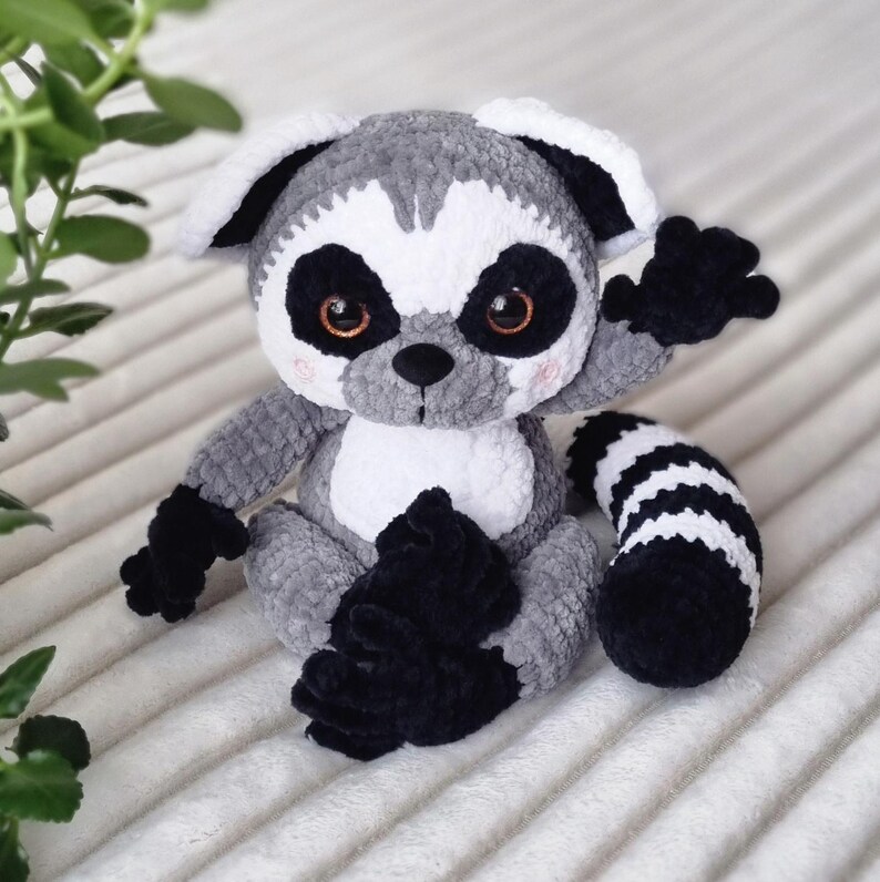May include: A gray and white crocheted lemur plush toy with black crocheted accents. The lemur is sitting with its arms raised and its tail curled behind it.