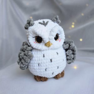 Könnte beinhalten: Eine handgefertigte, weiße Häkel-Eule mit grauen Akzenten. Die Eule hat große, ausdrucksstarke Augen mit goldenem Glitzer und rosigen Wangen. Die Flügel und Federohren sind grau, der Schnabel gelb. Die Eule ist ein weiches, plüschiges Spielzeug.