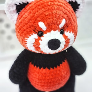 May include: A crocheted red panda plush toy with black and white details. The toy has a red body, black legs and arms, and a white face with black eyes and nose.