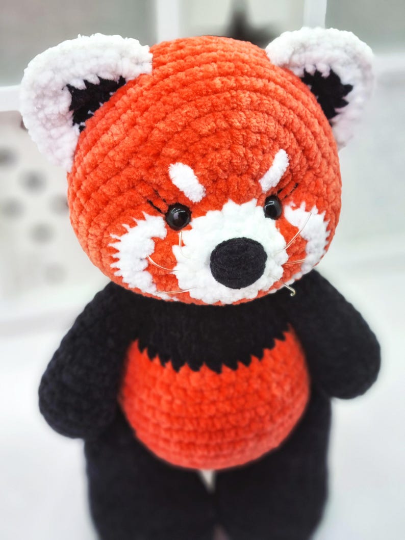 May include: A crocheted red panda plush toy with black and white details. The toy has a red body, black legs and arms, and a white face with black eyes and nose.