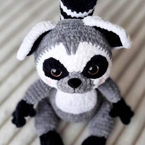 May include: A crocheted gray and white lemur plush toy with black eyes and a black tail. The lemur is sitting on a white and gray striped surface.