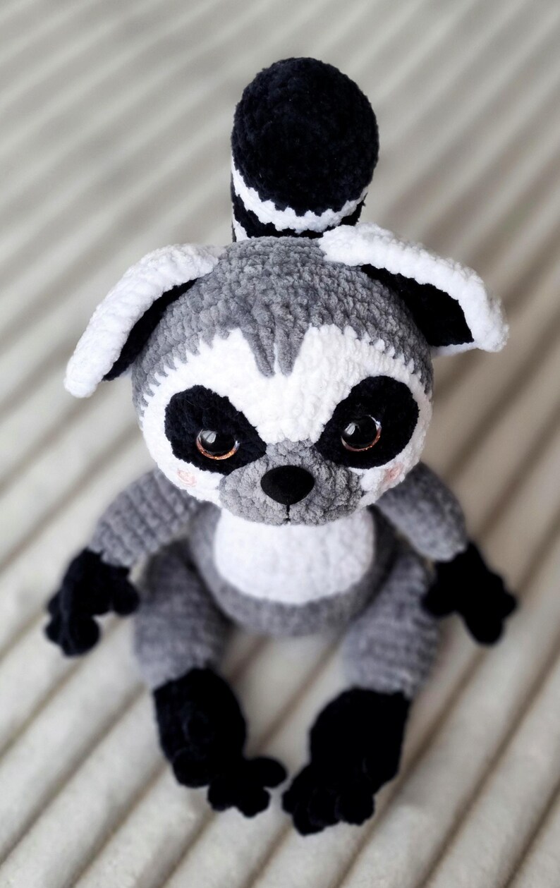May include: A crocheted gray and white lemur plush toy with black eyes and a black tail. The lemur is sitting on a white and gray striped surface.