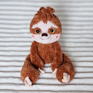 Pode incluir: Um brinquedo de pelúcia de preguiça de crochê marrom com um nariz rosa e grandes olhos marrons. A preguiça está sentada em uma superfície listrada branca e marrom.