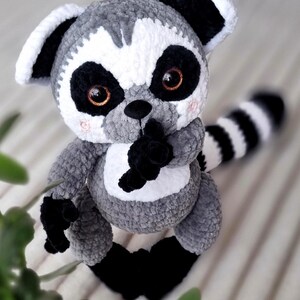 May include: A gray and white crocheted lemur plush toy with black eyes and a black and white striped tail. The lemur is sitting on a white and gray striped surface.