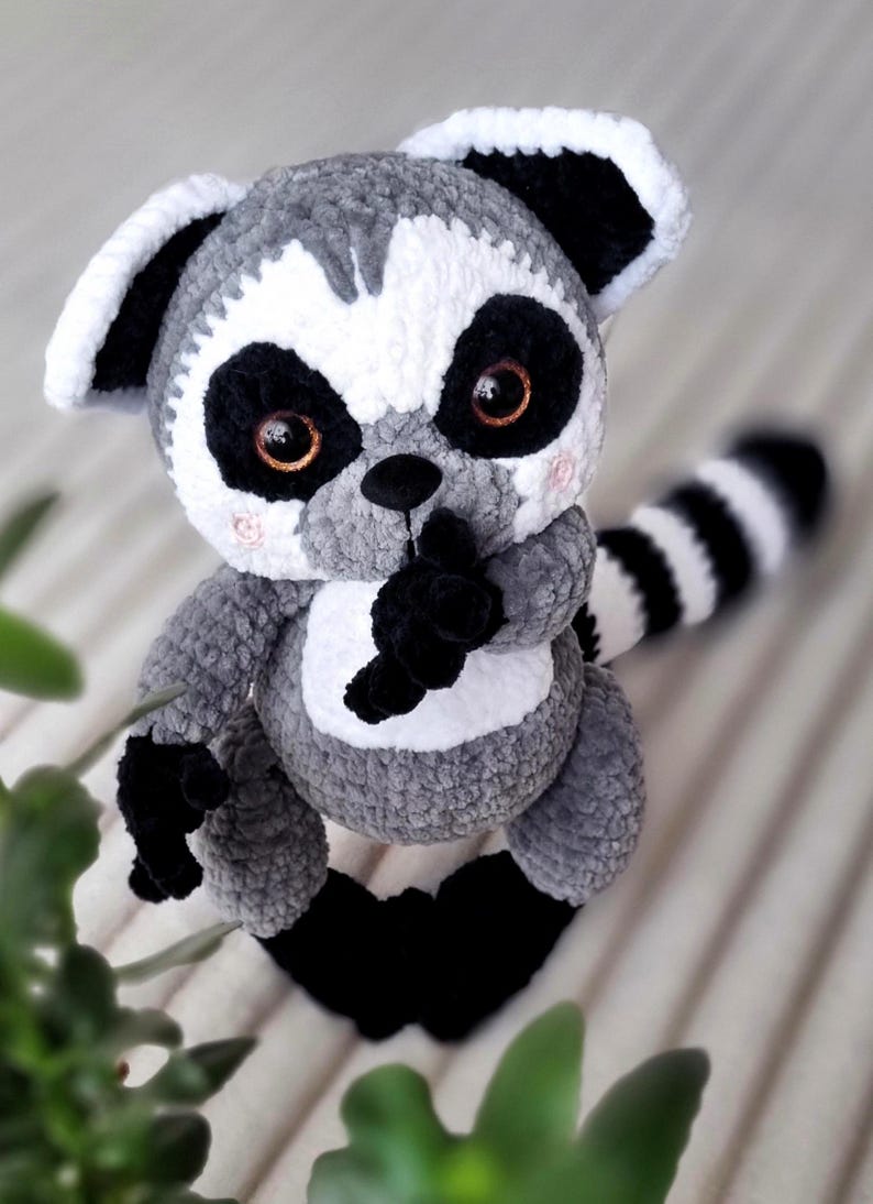 May include: A gray and white crocheted lemur plush toy with black eyes and a black and white striped tail. The lemur is sitting on a white and gray striped surface.