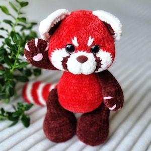 May include: A crocheted plush toy red panda, made with red and white yarn. It has brown legs and a white face with black eyes and a pink nose.