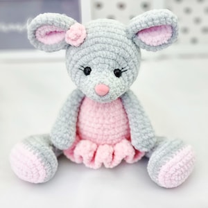 Puede incluir: Un ratón de crochet gris que lleva un vestido rosa con una flor en la cabeza. El ratón tiene ojos negros y una nariz rosa.