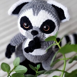 May include: A gray and white crocheted lemur plush toy with black eyes and a black nose. The lemur is sitting on a white surface with green leaves in the background.