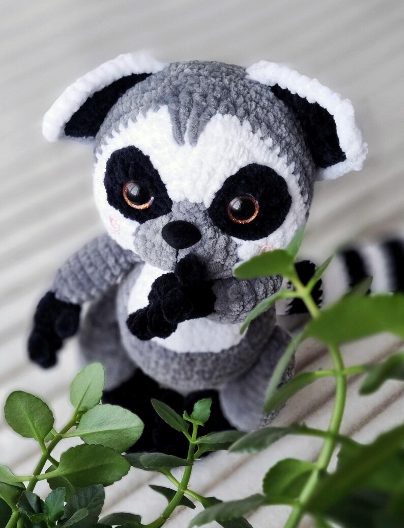 May include: A gray and white crocheted lemur plush toy with black eyes and a black nose. The lemur is sitting on a white surface with green leaves in the background.