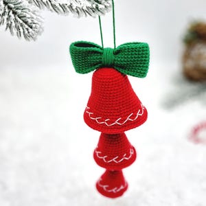 May include: A handmade Christmas ornament featuring three red crocheted bells with white stitching, topped with a green crocheted bow. The ornament is suspended from a green string, perfect for holiday decor.