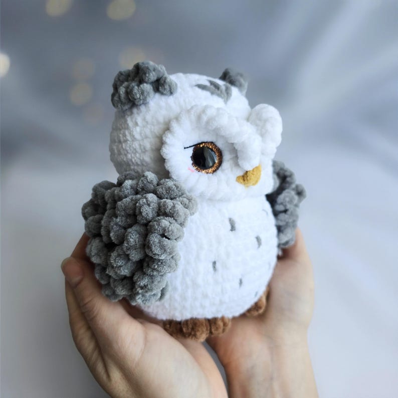 Pu&ograve; includere: Un gufo all'uncinetto bianco e grigio fatto a mano con piedi marroni e occhi bordati d'oro. Il gufo ha ciuffi grigi sulla testa e sulle ali. Il peluche &egrave; tenuto tra le mani di una persona.
