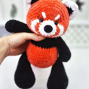 May include: A crocheted red panda plush toy with black legs and white accents around the face. The toy has black eyes and a black nose.
