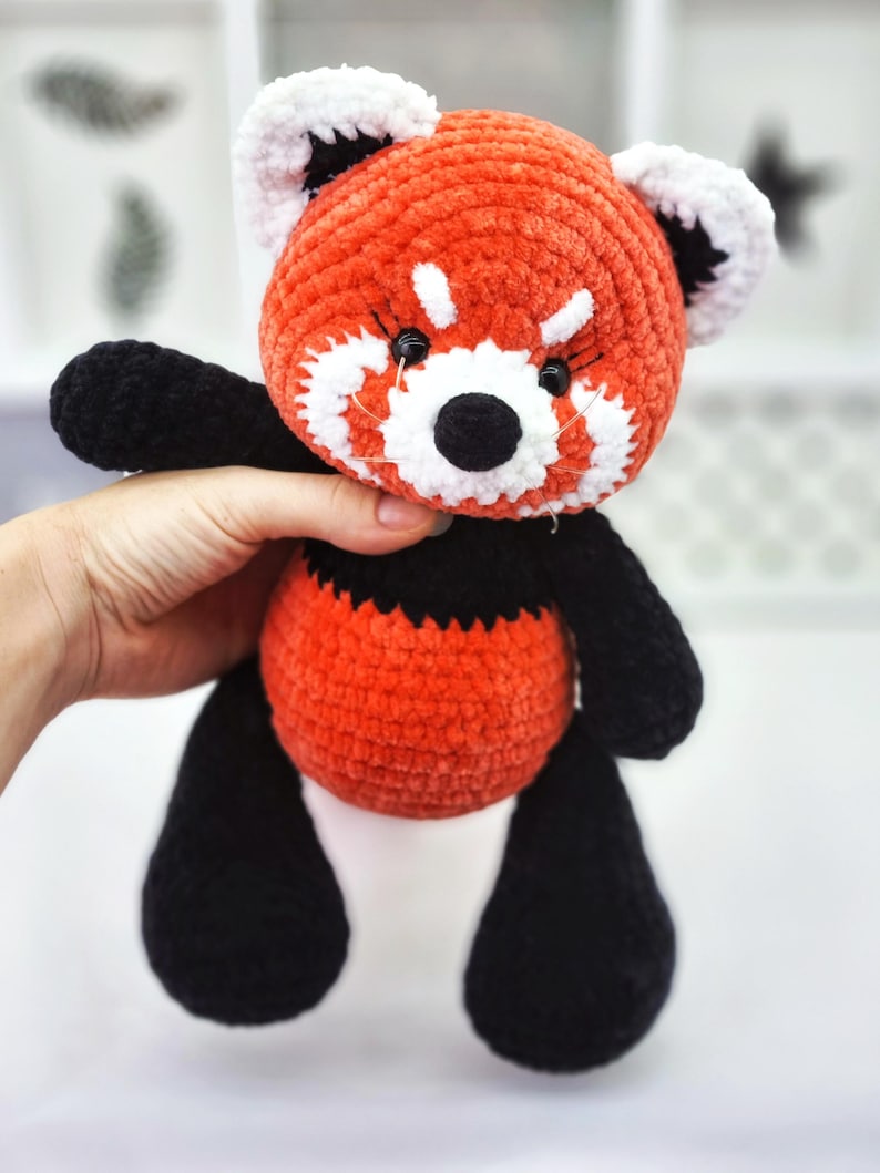 May include: A crocheted red panda plush toy with black legs and white accents around the face. The toy has black eyes and a black nose.