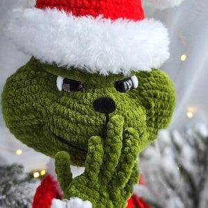 Peut inclure: Une figurine verte du Grinch en crochet, portant un bonnet de Père Noël rouge avec une bordure blanche. Le Grinch a une expression espiègle, une main couvrant sa bouche. Décoration de vacances festive.