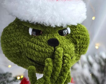May include: A green crocheted Grinch figure wearing a red Santa hat with white trim. The Grinch has a mischievous expression, with a hand covering its mouth. The figure is a festive holiday decoration.