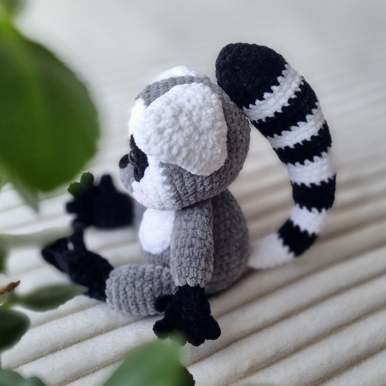 May include: A crocheted lemur toy with a long, striped tail. The lemur is sitting on a white surface and is made of gray, white, and black yarn.