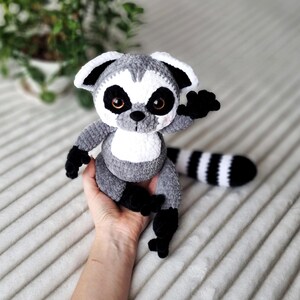 May include: A gray and white crocheted lemur plush toy with black stripes on its tail. The lemur is sitting on a hand and has a friendly expression.