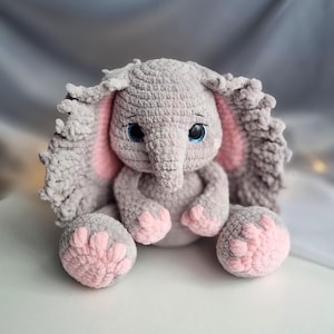 Puede incluir: Un juguete de peluche de elefante gris tejido a crochet con orejas y pies rosas. El elefante tiene ojos azules y una pequeña trompa.