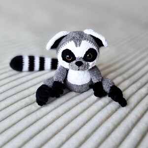 May include: A crocheted gray and white lemur plush toy with black eyes and a striped tail. The lemur is sitting on a white textured surface.