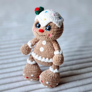 May include: A crocheted gingerbread man toy with white icing, colorful sprinkles, and a red and green candy on top. The toy has blue eyes, a red nose, and a white button on its chest.